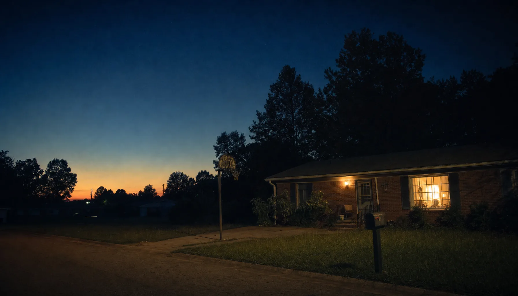 A brick ranch house at dusk with the porch light on and one window lit